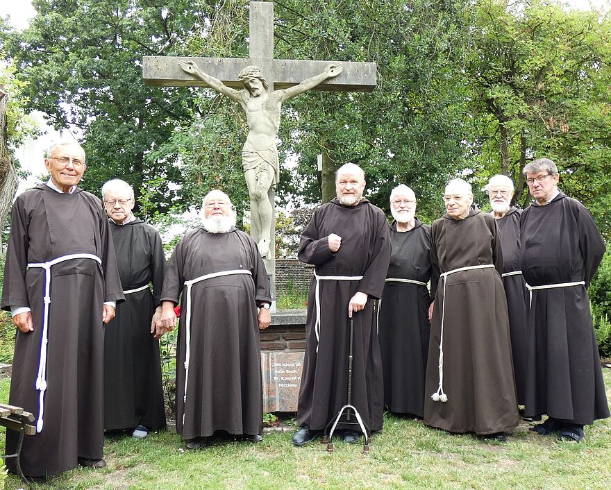 Acht Kapuzinerbrüder stehen in einem Garten vor einen steinernen Kreuz mit einer Christusfigur.